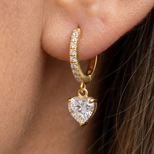 Gold hoop earring with a heart-shaped diamond pendant worn by a person.